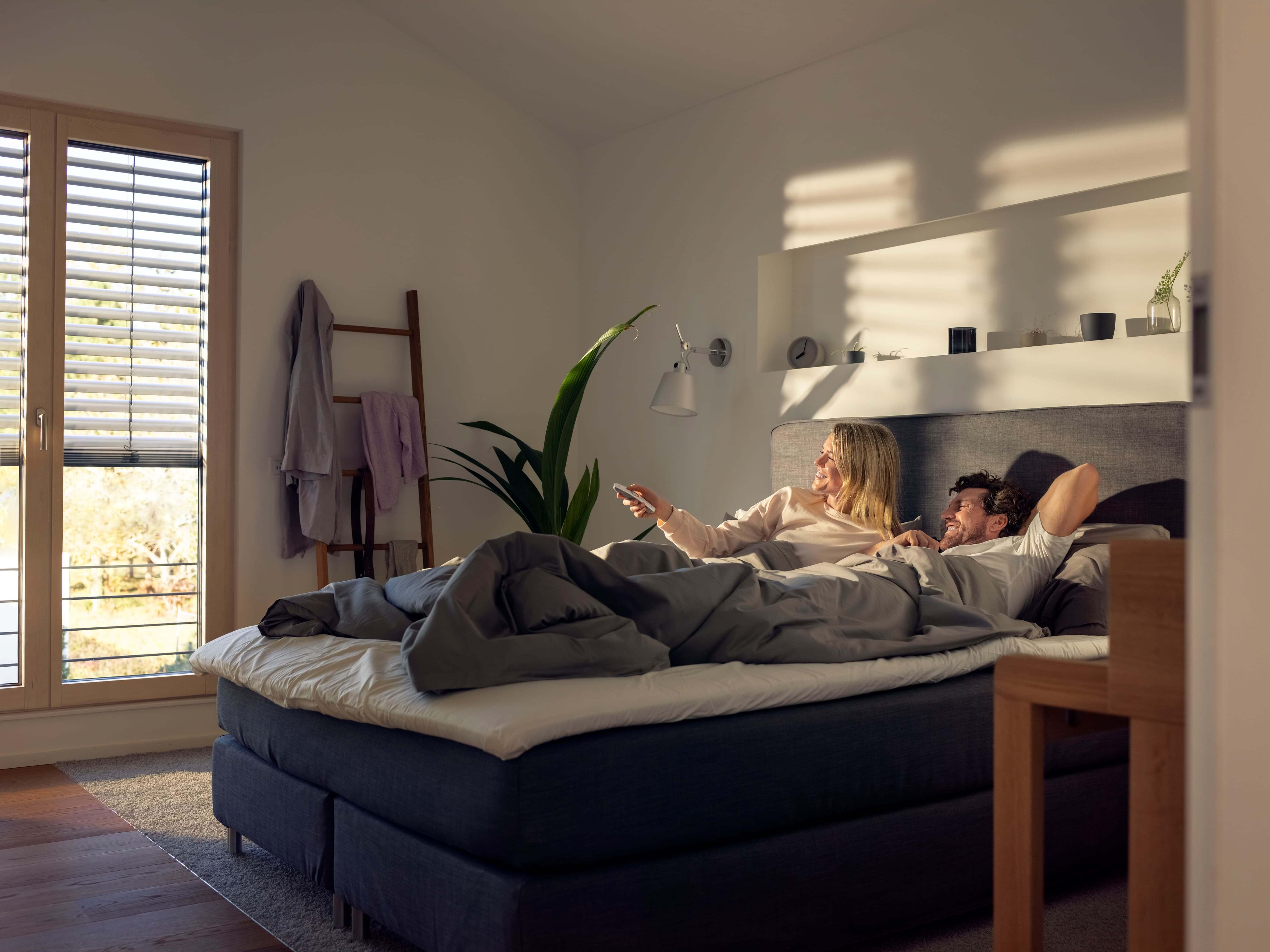 Ein Pärchen liegt im Bett in ihren Schlafzimmer und bedienen komfortabel ihren Sonnenschutz per Fernbedienung