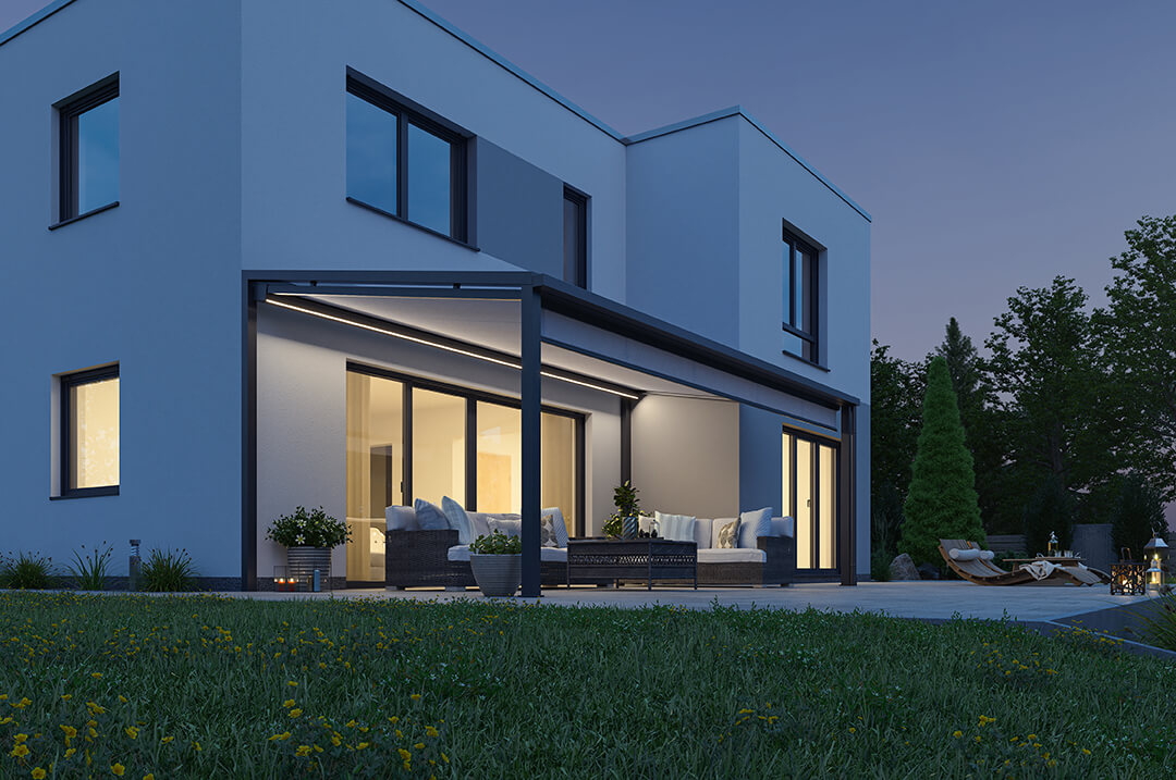 Terrasse eines Einfamilienhauses am Abend mit einem Terrassendach. Die Unterglasmarkise hat integrierte LED Beleuchtung im Kasten und in den Führungsschienen und leuchtet die Terrasse schön aus.