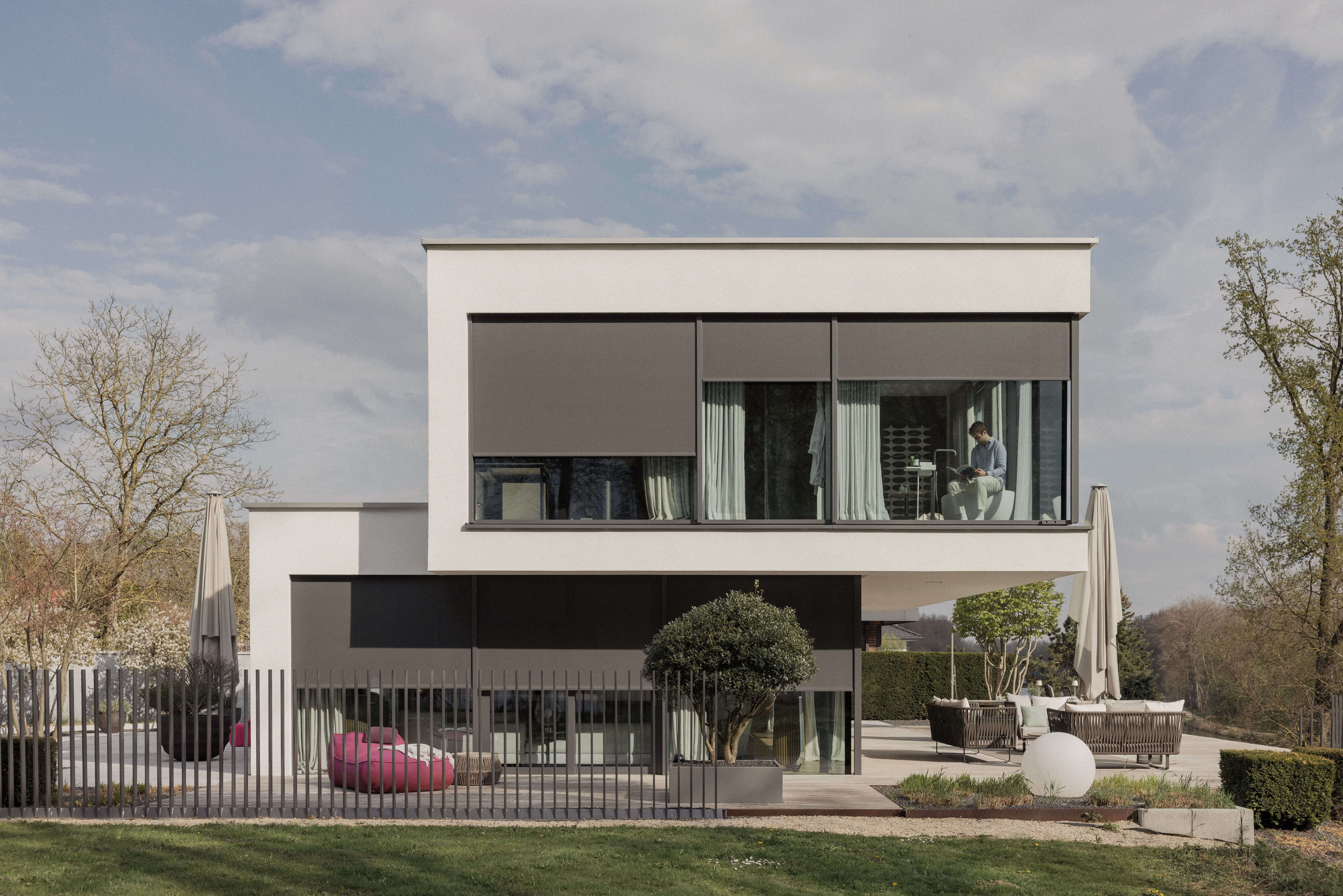 Sehr modernes kubisches Einfamilienhaus vom Garten ausgesehen. An den großen Glasfronten sind Textilscreen heruntergelassen und den Innenraum zu schützen.