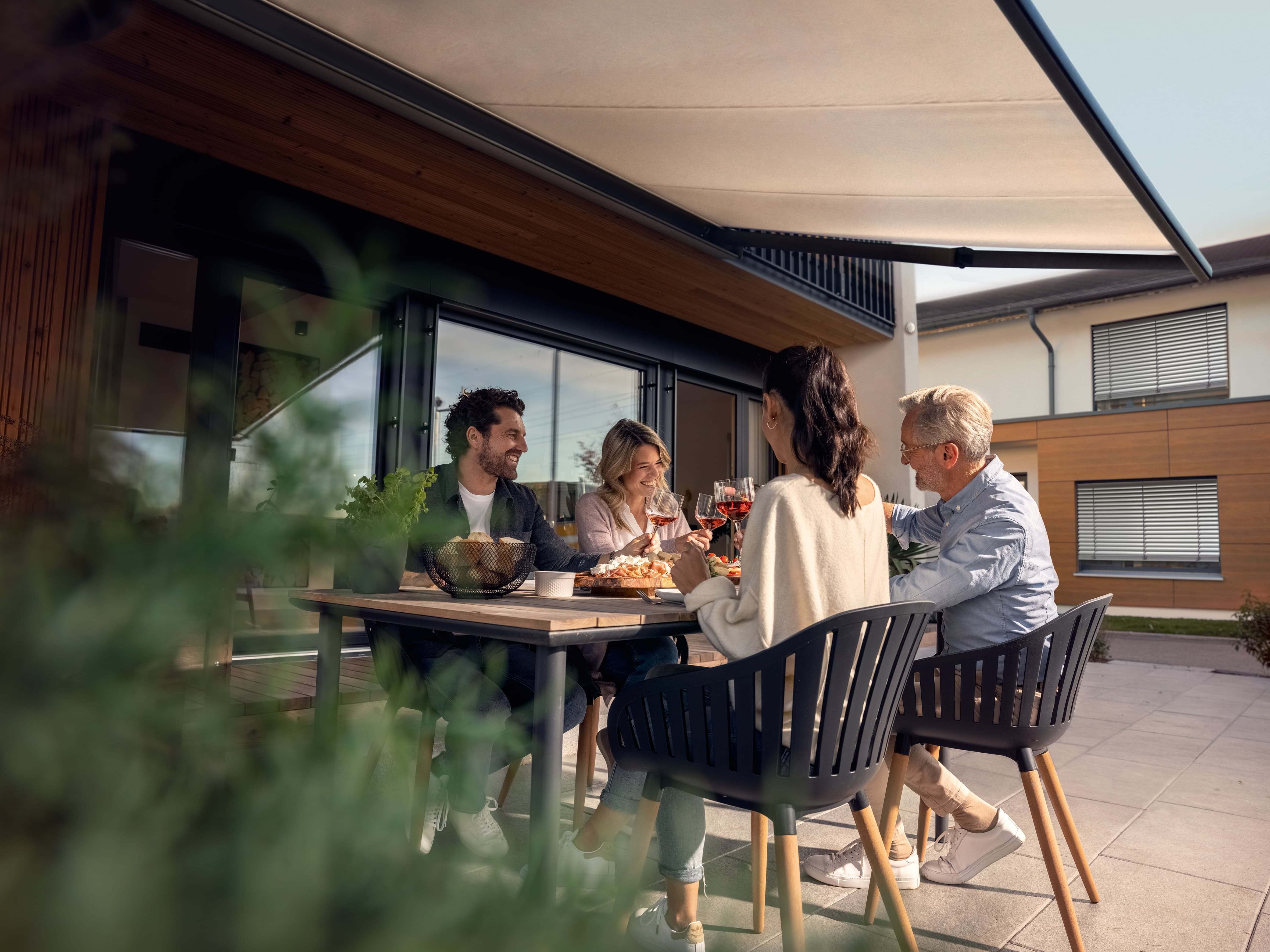 Eine Terrasse auf der vier Personen an einem Tisch sitzen. Oben ist eine ausgefahrene Markise, die die Leute vor der Sonne schützt.